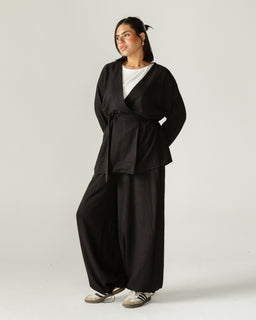 A black wrap-and-flow set featuring a long-sleeve, V-neck wrap top with a matching wide-leg pant. The outfit appears designed for women, with a relaxed, draped silhouette and a tie belt at the waist. Notable materials seem to be a soft, matte fabric in solid black, emphasizing a loose, comfortable, lounge-to-everyday style.