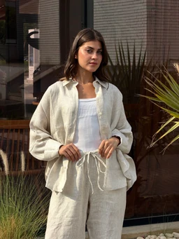 - What the product is: A beige linen round-neck shirt set with matching drawstring waist shorts, worn as a coordinated loungewear outfit.
- Intended audience: Unisex-looking style suitable for women or girls based on the model and proportions; presented as comfortable, casual loungewear.
- Notable attributes visible: Natural beige linen fabric with a relaxed fit, long-sleeve shirt layered over a white tank, and a pair of matching linen shorts with a drawstring tie. The look emphasizes a soft, airy texture and casual, oversized silhouette.
