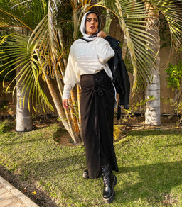- What it is: A women’s outfit featuring a white hoodie/sweater paired with a high-waisted black wrap-skirt, styled with a black jacket draped over the shoulder.
- Intended audience: Women (adult).
- Notable attributes: White long-sleeve top, black wide-leg wrap skirt with a front knot detail, black combat boots, black outerwear draped over the shoulder, and a light headscarf. Material impression: soft knit top and flowy skirt fabric; color palette is white and black. Style: casual, modest, streetwear-inspired.