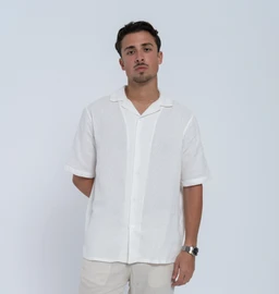 - A lightweight short-sleeve button-front shirt in solid white, worn by a male model.
- Appears styled for warm weather with a relaxed, casual cut and a camp/cuban-style collar.
- Notable attributes: soft, breathable fabric with a relaxed drape; short sleeves; front placket with visible vertical seam; clean white color; slightly longer length that sits over the hips. Audience: men. Material hints: linen or linen-blend feel due to texture and drape. Style: casual/laid-back shirt.