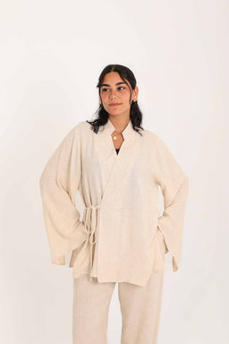 - A beige linen blouse with a loose, draped silhouette and wide, kimono-style sleeves. It features a lightweight, natural-textured fabric and a tie belt at the waist, worn over matching beige pants in a relaxed, coordinated set.
- Target audience: women, based on the cut, styling, and draped femininity of the garment.
- Notable attributes: linen material, beige/cream color, long flowy sleeves, V-neck with wrap-style front, waist tie detail, and a relaxed, oversized-fit top paired with matching wide-leg pants.