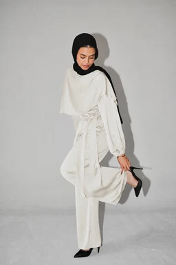 - This image shows a white satin wrap set worn by a female model. The outfit appears loose and flowing with a draped wrap over a long-sleeve top and wide-leg pants, all in ivory satin.
- Intended audience: women's size and styling, suggested by feminine silhouette, hijab styling, and the soft, flowing fabric.
- Notable attributes visible: white satin fabric, wrap design with a sash/belt at the waist, long sleeves with cuffs, loose cape-like overlay or shawl over the shoulders, and black headscarf and black shoes for contrast. The look is elegant, minimal, and monochrome.