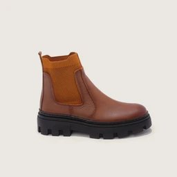 A brown Chelsea-style ankle boot with a chunky black rubber sole. It features a smooth leather upper, elastic side panels in orange-brown knit, and pull-tab at the back for easy on/off, giving a rugged, utilitarian look. The boot appears designed for men or unisex wear, with a substantial tread and durable construction.