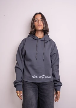 - A cropped, long-sleeve hoodie in a muted gray/blue tone with a drawstring hood and relaxed fit. The model is a woman wearing dark wash jeans to pair the cropped hoodie, suggesting a casual, streetwear aesthetic for women. Notable details include the visible white script text “nice not naïve” printed at the front hem, and a soft, fleece-like material typical of casual hoodies.