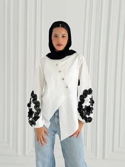 - Description: A white buttoned chemise with an asymmetric wrap silhouette and long sleeves. The garment features diagonal front buttons and black embroidered floral appliqué along the outer sleeves near the cuffs.
- Intended audience: Women's.
- Notable details: Off-white/white fabric, black embroidery on sleeves, voluminous sleeves with gathered cuffs, relaxed-fit with an asymmetrical buttoning and wrap design. Material appears lightweight cotton or cotton-blend. The model wears a black headscarf for styling in the photo.