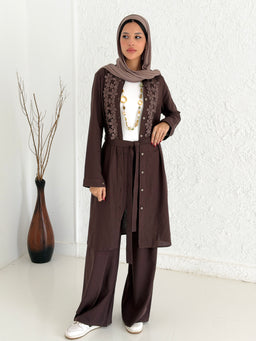 This is a modest, brown linen shirt set with a long tunic and matching wide-leg pants, designed for women. The model wears a light beige hijab and a white inner top with a gold-toned necklace, giving a layered, traditional-urban look. Notable details include the long-sleeve tunic with embroidered panels along the front, a self-tabric belt/closure, and the soft, matte brown linen fabric in a relaxed silhouette.