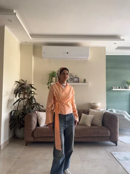 - What the product is: A wrap-style shirt in orange, designed to be tied at the waist for a wrap silhouette.
- Apparent audience: Women (model appears female; styling with a wrap front, long sleeves, and scarf/neck accessory suggests a women's fit).
- Notable visible attributes: Bright orange fabric, long sleeves with a wrap front and tie belt extending as a sash, lightweight and slightly draped; paired with blue jeans in the photo. The setting shows an indoor living room with neutral-toned decor.