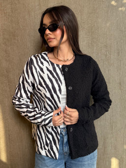 - What the product is: A zipped or button-front cardigan/jacket featuring a two-tone color block design. One half is zebra-print black-and-white, the other half solid black with a textured knit.
- Apparent audience: Women's (model styling, slim fit, over a casual outfit with sunglasses and accessories).
- Notable attributes: Black and white zebra-print panel paired with a solid black textured knit panel; mid-weight cardigan with buttons down the front; cropped to hip length; long sleeves. Material appears to be a fuzzy, bouclé or boucle-like knit.