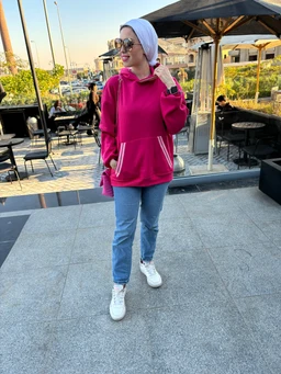 - What the product is: A pink hoodie with a front pocket and contrasting white trim detailing, shown being worn as a casual outer layer.
- Apparent audience: Women (based on fit, styling, and model).
- Notable attributes: Bright pink color, relaxed pullover cut, front kangaroo pocket with light-colored trim, long sleeves, hood, and white sneakers paired for a casual look. Material appears to be a soft sweatshirt fabric. The hoodie is styled with light blue jeans and a white headscarf in an outdoor setting.