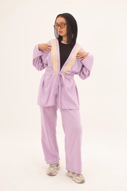 - Product: A lilac-colored plush loungewear set consisting of a robe-style cardigan with a belt and matching wide-leg pants.
- Intended audience: Women (based on styling and fit with the model).
- Notable attributes: Soft, fuzzy lilac fabric; cream/white trim along the collar and lapels; belt-tie at the waist; black top worn underneath; relaxed, oversized sleeves; light-colored chunky sneakers.