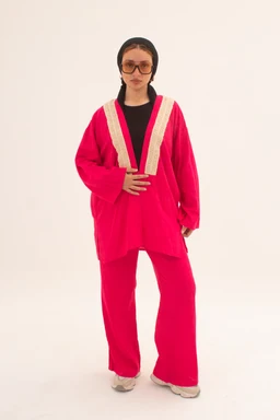 - What the product is: A bright pink knit cardigan with a loose, oversized fit paired with matching wide-leg pants; it features a deep V-neckline trimmed with beige crochet or knit detailing.
- Intended audience: Women (judging by the styling, oversized silhouette, and facial presentation; the look is feminine and contemporary).
- Notable attributes: Hot pink fuchsia color, beige contrast trim along the neckline, long balloon sleeves, relaxed cropped-length cardigan over wide-leg pants, light-colored (cream/white) shoes, and a dark inner top. The overall style is bold, cozy, and retro-inspired.