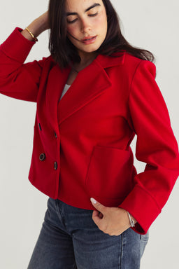 A red structured blazer-style jacket with a double-breasted front and wide notched lapels. Appears tailored for women, shown on a female model with mid-length dark hair and a casual pose, suggesting a smart-casual or office-ready look. Notable features include bold red fabric, black metal button accents, slightly cropped length, and long sleeves. Visible material looks like a medium-weight woven fabric with a crisp finish.