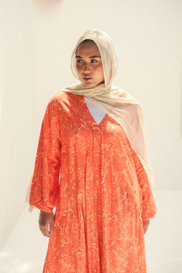 A loose, ankle- or knee-length abaya or dress in bright orange with a delicate light pattern. It’s styled for women, shown with a light beige hijab and a white inner top, giving a modest, flowing silhouette. Notable attributes include the soft, sheer fabric, wide 3/4 sleeves, V-neckline, and the warm orange color with subtle botanical-like pattern.