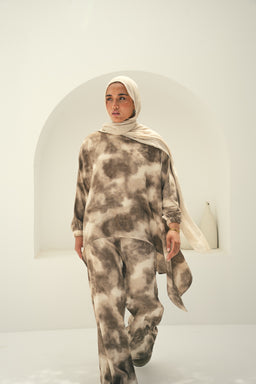 - A modest, relaxed-fit loungewear set consisting of a tie-dye print long-sleeve top and matching wide-leg pants. The model is wearing a light-colored hijab, suggesting the outfit is targeted to women. The fabric appears soft and drapey, with a neutral taupe and beige color palette. The look is finished with a flowing scarf-like accessory draped over one shoulder, giving a comfortable, casual, modest style.