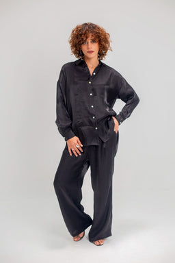 - Product: A shimmery black pajama set with a long-sleeve button-up shirt and wide-leg pants. The fabric appears silky and glossy.
- Intended audience: Women (based on the model, styling, and cut).
- Notable attributes: Dark black/sleek shimmery finish, relaxed fit, long-sleeve button-down shirt with a collar, matching wide-leg pants, and open-toe sandals shown.