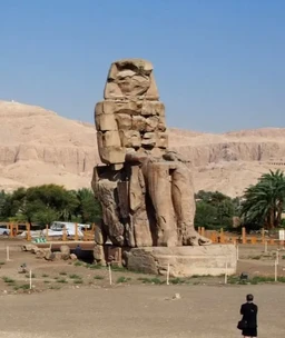 - What the product is: A travel or tour photography/advertisement image featuring an ancient Egyptian statue site with sandstone obelisk remains and desert surroundings.
- Apparent intended audience: Unisex; suitable for travel enthusiasts and history lovers viewing a day-tour to Luxor.
- Notable visible attributes: A large stone statue/ruin in a desert setting, with palm trees and low fencing in the foreground, clear blue sky, and a person in dark clothing walking near the site. Earthy beige-brown tones dominate; no branding visible.