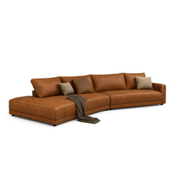 A large sectional sofa in warm cognac brown leather with a low-profile, modular layout. It appears designed for a contemporary living room, likely shown as unisex appeal due to neutral color and sleek, modern lines. Notable materials and details include smooth cowhide leather upholstery, plush seat and back cushions, and a long chaise segment with a soft, draped throw and assorted throw pillows for contrast.