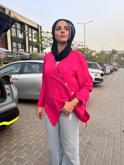 - Item: A bright pink cardigan-style blouse with a wrap/hem detail and wooden-tone buttons across the front.
- Audience and styling cues: Appears tailored for women, modeled with a black headscarf and casual streetwear vibe, suggesting a unisex-friendly, modest styling for everyday wear.
- Materials and features: Knit fabric with a chunky rib texture, long sleeves, asymmetrical hem, and visible wooden buttons; paired here with light gray wide-leg pants for a relaxed, casual look. Color is vivid fuchsia/pink.