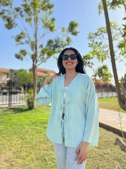 - A mint green tie-front top with a loose, relaxed fit, modeled as a casual outer layer over a light top.
- Intended audience appears to be women, based on the styling, cut, and feminine silhouette.
- Notable attributes: pastel mint color, long sleeves, front tie closures with multiple tie sections, lightweight fabric, with a soft, casual, breezy vibe. The look is styled with light denim or bottom wear and dark sunglasses in an outdoor park setting.