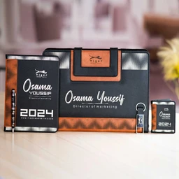 - Product: A matching travel/office set including a black and brown two-tone organizer or briefcase with a front logo panel, a striped black-and-brown notebook, and two small card-sized items. 
- Audience: Unisex (suitable for both men and women) based on neutral styling, compact size, and professional vibe.
- Notable attributes: Black and brown color scheme with tan accents; material appears to be faux leather or coated fabric with metal hardware; includes a large zippered case, a slim notebook, a small card-holder or USB-like item, and a mini card or file holder. The styling is modern and professional, suitable for marketing or office use.