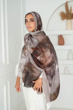 - What the product is: A printed cotton scarf styled on a model, shown draped over the head and shoulders with one end wrapped loosely at the chest.
- Apparent audience: Unisex or women’s accessory (model styling and hijab-friendly presentation suggest women; appears suitable for all genders who wear headscarves).
- Notable attributes: Soft, breathable cotton with an earthy brown, taupe, and white abstract print. Light to mid-weight scarf with a slightly sheer, flowing drape and finished edges.