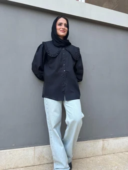 - What the product is: A black button-front shirt with a large, decorative collar and shoulder details.
- Intended audience: Women (model wearing it appears feminine; hijab styled, long sleeves, tailored fit).
- Notable attributes: Solid black fabric with long sleeves, exaggerated scalloped/ruffled bib collar extending over the shoulders, front placket with visible buttons, relaxed yet structured fit, paired here with light-wash jeans. Material appears lightweight woven.