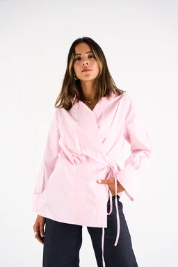This image shows a women’s fashion top styled as a tie-front shirt or blouse in baby pink. The model appears to be adult female; the cut suggests a relaxed, wrap-style shirt with a waist tie, long sleeves, and a slightly oversized fit. The fabric looks crisp and lightweight, with a soft pink hue and a casual, day-to-night aesthetic. The shirt is paired with dark jeans, highlighting a versatile, chic casual look.