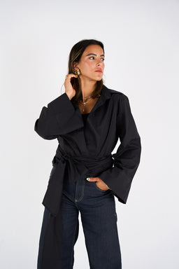 - What the product is: A black wrap shirt with a tie-front detail and wide, voluminous sleeves styled on a casual-wardrobe look. The wrap creates a cinched waist silhouette.
- Apparent audience: Women, based on the fit, sleeves, and styling.
- Notable attributes visible: solid black color, lightweight woven fabric, oversized collar, long bell/kimono-inspired sleeves, wrap-around waist tie, cropped-ish hem, paired with dark jeans. No brand visible.