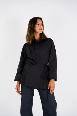 This is a black, long-sleeve shirt/jacket with a wrap-style front and a self-tabric belt at the waist, creating a structured silhouette. The sleeves are wide with turned-up cuffs, and the length falls at hip level. The styling suggests a versatile, smart-casual piece likely aimed at adults (women) for dressing up or down, with a lightweight, matte fabric and a tailored, modern look. Notable attributes: black color, wrap-waist belt, cuffed rolled sleeves, and a clean, minimal design.