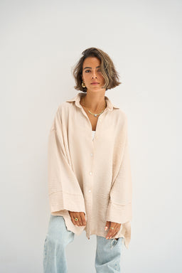 The Go-To Oversized Shirt in a light floral white appears as a loose, relaxed-fit button-up blouse with a soft, airy drape. The model’s styling suggests a unisex or women’s silhouette, with an oversized, longline shape and dropped shoulders, paired here with light denim for a casual look. Notable attributes include the creamy white fabric, a subtle floral pattern or texture, a classic collar, and a front placket with visible buttons.