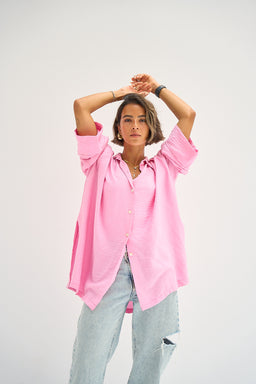 A loose-fitting oversized shirt in a soft blush pink, worn open over a simple base layer. Designed for a unisex or relaxed women’s fit, with dropped shoulders and long sleeves that can be rolled or scrunched for styling. Visible features include a button-front placket, a dipped hem, and a lightweight cotton-like fabric with a casual, breezy silhouette.