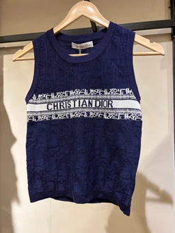 What it is: A sleeveless knit tank top with a high crew neck, presented on a wooden hanger.
Intended audience: Women (feminine styling, cropped armholes and curved hem).
Notable attributes: Dark navy blue lace-knit body with a white embroidered/knit band across the chest featuring repeating pattern and the text “CHRISTIAN DIOR” in black letters. Lightweight knit fabric with a fitted silhouette, ribbed neckline and armholes.