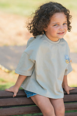 - What the product is: A loose, oversized short-sleeve tee with a relaxed fit.
- Intended audience from visible model: Children's (kid) sizing, shown on a young child.
- Notable attributes: Light gray-beige color, short sleeves, crew neckline, small floral embroidery on the chest, paired with denim shorts. The fabric appears soft and casual with a roomy silhouette.