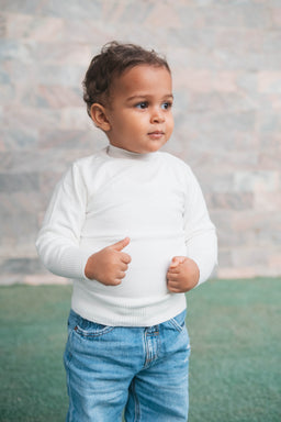 - Product: White turtle neck sweater for babies. The photo shows a young child wearing a plain white knit turtleneck with long sleeves and a ribbed hem, paired with blue jeans. 
- Intended audience: Baby/infant (unisex style appears suitable for both boys and girls based on the neutral white color and simple design).
- Notable attributes: White knit fabric, long sleeves, high turtleneck neckline, ribbed cuffs and hem, casual everyday styling.