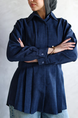 - What the product is: A dark blue, long-sleeve button-up shirt with a loose, pleated peplum-style cut.
- Apparent intended audience: Women (based on the styling, fit, and the model’s presentation).
- Notable attributes: Deep navy color, textured woven fabric, collared neckline, long sleeves with cuff buttons, and a flared, pleated hem creating a relaxed yet structured silhouette. The model is wearing a headscarf, suggesting modest styling.