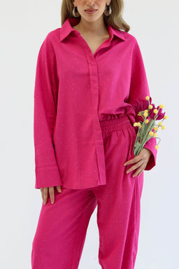 - What it is: A matching two-piece pajama set in a bright pink color, featuring a long-sleeve button-front shirt and elastic-waist pants.
- Audience and styling cues: Designed for adults; feminine styling evident in the relaxed fit, soft drape, and delicate dotted texture; the model holds a small bouquet, emphasizing a soft, lounge-to-casual vibe.
- Visible materials and details: Bright pink fabric with a subtle dotted pattern; collared shirt with a placket, dropped shoulders, and full sleeves; elastic waistband on the pants; relaxed, straight leg silhouette. No visible branding.