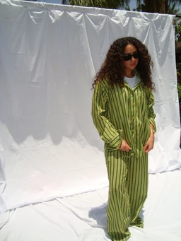 - Description: A model wearing a matching green striped loungewear set consisting of a long-sleeve button-up shirt and wide-leg pants. The outfit is loose-fitting and casual, with vertical olive-green and darker green stripes.
- Audience and styling: Appears designed for women (feminine fit, relaxed styling) suitable for lounge or casual wear.
- Materials and colors: Primary color is olive/green with darker green stripes; fabric looks lightweight and soft, like cotton or a jersey knit. The look is laid-back and streetwear-influenced, complemented by dark sunglasses.