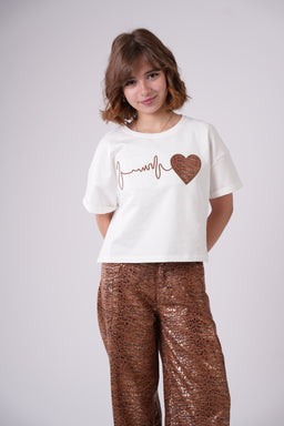 - What the product is: A white cropped graphic tee with a heart-themed design, modeled by a young girl.
- Apparent audience: Girls (youth/child sizing), based on the model and styling.
- Notable attributes: Short-sleeve white knit top with a brown graphic line and heart motif across the chest; paired with bronze sequin pants in the photo. The shirt has a relaxed, cropped fit and a crew neckline.
