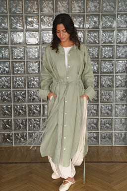 - What the product is: A long, lightweight shirt-dress made from pure linen, shown in a sage green color with a white inner layer peeking at the neckline and hem. It features a button-front closure, tie-waist drawstring, and split hand-access pockets, styled with a relaxed, oversized fit and long sleeves gathered at the cuffs.
- Apparent audience: Women (styled on a female model with a feminine, relaxed silhouette).
- Notable attributes: Pure linen fabric in sage green with a white underlayer, button-down front, drawstring waist, mid-calf to ankle length with a curved hem, cuffed sleeves, and casual, breathable styling.