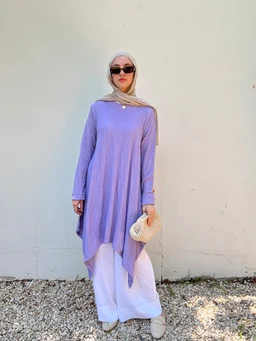 - What the product is: A crinkle, high-low knit top in a soft purple-lavender shade, styled over wide-leg white pants.
- Intended audience: Women (modest, hijab-friendly styling with a headscarf and sunglasses signals a female audience).
- Notable visible attributes: Long-sleeve tunic with a high-low hem, crinkled texture, relaxed fit, paired with white wide-leg pants; soft lilac-purple color; paired with a headscarf, oversized sunglasses, and a small woven bag for a casual modest look.