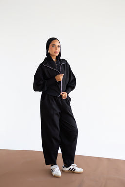 - What it is: A black zip-front hooded windbreaker paired with loose-fitting black wide-leg pants. The model is wearing white sneakers with black stripes.
- Audience and styling cues: Appears as unisex or women's casual athleisure; relaxed fit with a slightly oversized silhouette, cropped jacket length, and roomy pants.
- Materials, color, and notable attributes: Black fabric with a subtle sheen suggests lightweight synthetic or windbreaker material; white zipper and white sneakers create contrast; overall minimalist streetwear style.