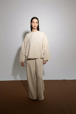 - A model wears an oversized, monochrome beige loungewear set consisting of a long-sleeve top with dropped shoulders and wide, flared sleeves, paired with matching wide-leg pants.
- Target audience appears to adults (unisex-leaning but styled in a way that could be for women), based on the soft drape, relaxed fit, and neutral color. Notable attributes: modal fabric feel suggested by the name, neutral cream-beige colour, relaxed oversized silhouette, and minimalistic styling.