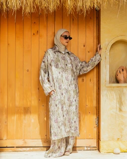 - The image shows a woman wearing a matching modest set: a long-sleeve, knee-length tunic with matching wide-leg pants. The outfit has a loose, relaxed fit and an all-over floral/abstract print in light, muted tones.
- Intended audience appears to be women (modest fashion styling, hijab, sunglasses). Notable attributes: soft, breathable fabric in a light, creamy-beige base with a delicate floral print; collared neckline with a buttoned placket; coordinated tunic and pants set. The look is finished with a hijab and sunglasses for a chic, casual-wleet style.