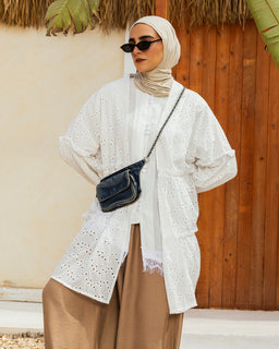 - What the product is: A long, white eyelet kimono-style outerwear piece styled over a white inner top, paired with a beige skirt and a small crossbody bag. The garment features oversized, voluminous sleeves and a knee-to-thigh length with an open front.
- Apparent audience: Women (based on hijab styling and feminine silhouette).
- Notable attributes: White eyelet fabric with a delicate cut-out pattern, oversized puff sleeves, relaxed open-front cardigan/kimono design, knee-length with light layering underneath, beige or nude inner clothing, black crossbody bag, and a light beige hijab. The overall look is modest, chic, and summery.