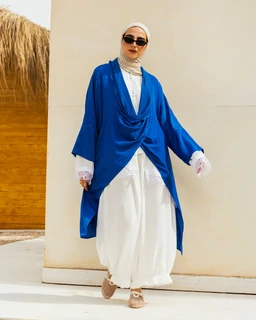 - What the product is: A modest outer layer over a white abaya-style outfit. It appears as a loose, draped open-front kimono/overlay in a vivid royal blue over a white inner garment.
- Target audience: Women (styled with a hijab and feminine drape, long sleeves and flowing silhouette).
- Notable attributes visible: bright blue satin-like drape with wide, flowing sleeves, layered over white long-sleeved inner garments and a white hijab. White trousers/pants with a soft, billowy silhouette and light sandals complete the look. No branding visible.