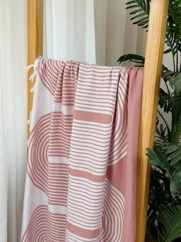 This is a soft, pink-toned towel or throw draped over a wooden stand. It appears to be unisex in styling, with a geometric pattern of curved and linear stripes in muted rose and white. The visible material looks like a plush cotton or terry fabric, with a faded-block color palette and tassel fringe along one edge.