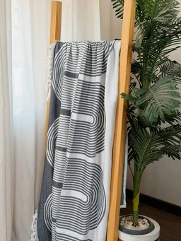 - What the product is: A woven textile accessory or cover, shown draped over a wooden frame with a geometric pattern.
- Apparent intended audience: Unisex (displayed on a neutral setup; no gendered styling cues).
- Notable attributes: Black and light gray geometric circular/curved stripe pattern on a light background; appears to be a soft, woven fabric with tassels at the edges. The photo presents it as a rectangular throw or towel-like fabric, shown in a home decor setting.