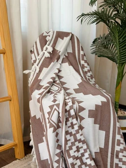 - Product: A cozy throw blanket draped over a chair, with white and brown geometric patterns and tassels along the edges.
- Audience: Unisex (suitable for both men and women) judging from neutral colors and cozy styling.
- Notable attributes: Thick knit/woven fabric, brown and white color scheme, bold geometric motifs, fringed tassels at the edges, draped over a chair in a home setting.