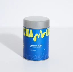 A small cylindrical tin container with a metallic lid, labeled Ceremonial Blend Matcha. It appears gender-neutral in styling, suitable for adults. The visible design features a bright blue body with yellow graphic accents and white text, suggesting a premium matcha powder product.