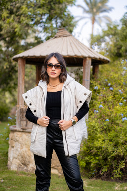 - A light-colored puffer vest with a dropped shoulder silhouette worn over a black top and black faux leather pants. The vest features a high collar and zip/knit closures with visible snap or button detailing on the sides, giving a padded, quilted look.
- Intended audience: women's (based on styling, fit, and model).
- Notable attributes: light beige/cream color, puffer quilting, oversized/dropped-shoulder design, sleeveless vest layered over a black long-sleeve top and glossy black pants, casual outdoor setting.